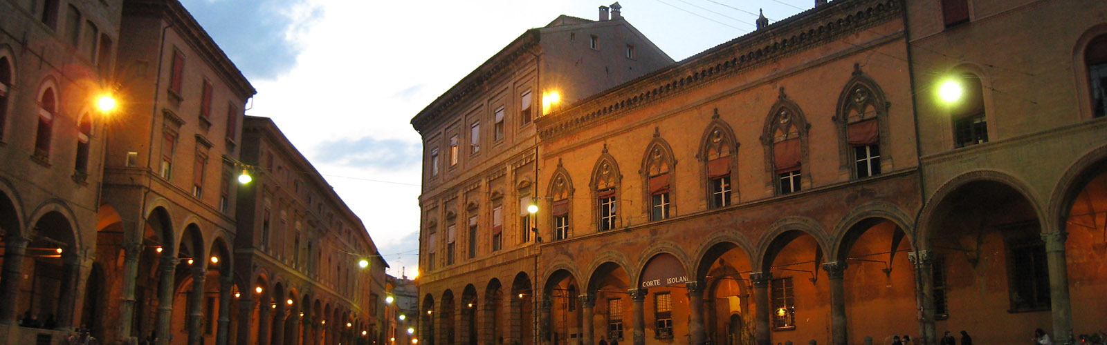 Two buildings, made out of limestone with several arches, ornate details, and windows, stand across from each other. There are many golden lights attached to the buildings glowing brightly.